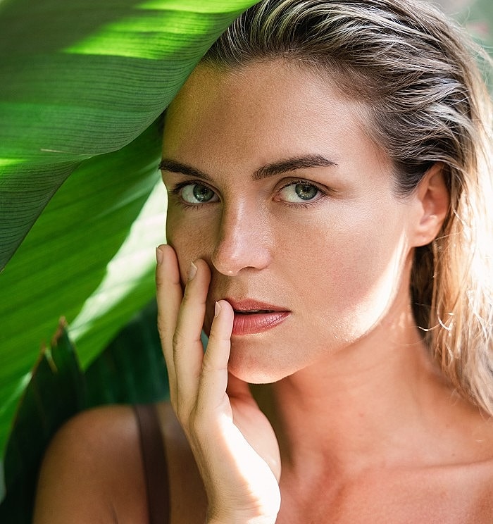 Woman with hand on face among green foliage.